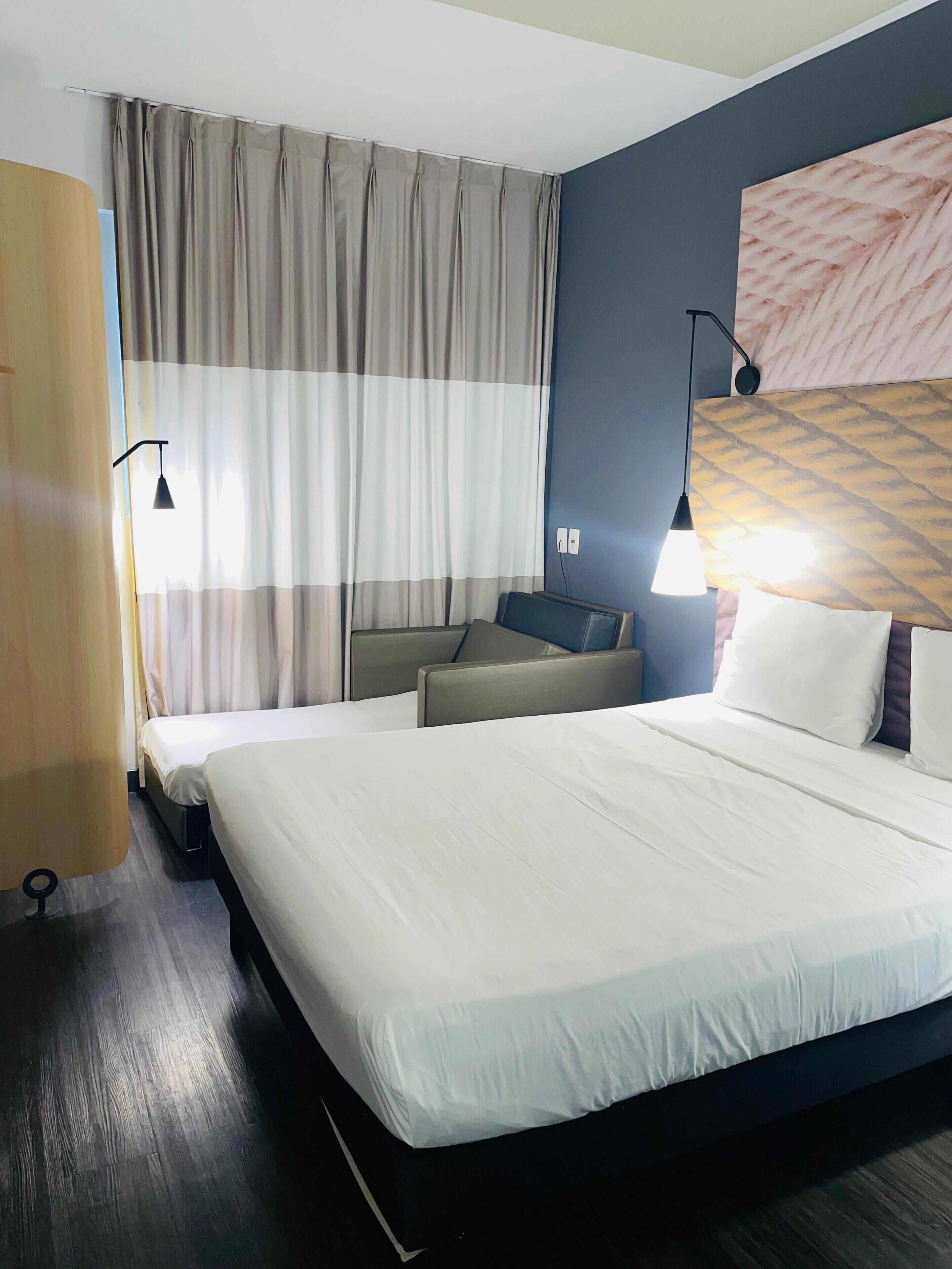 A hotel room with a large bed, two bedside tables, a headboard with a patterned design, a window with curtains, and a wall-mounted lamp.