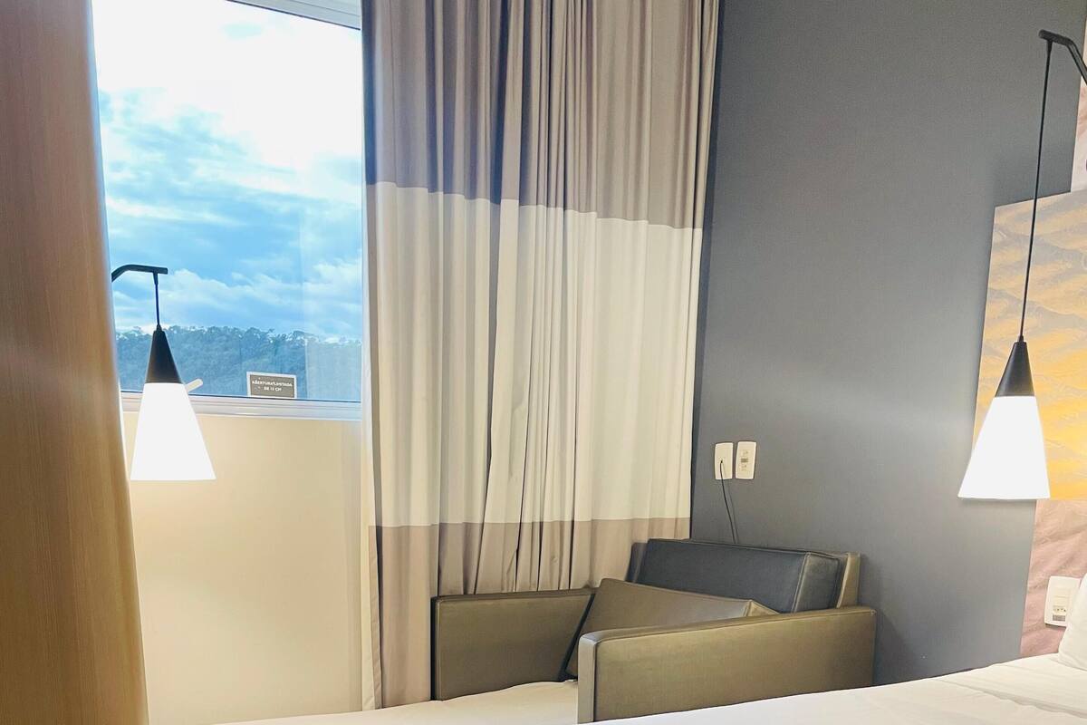 Habitación de hotel con una cama grande, un sofá y una mesita. Hay cortinas y una ventana con vistas al cielo.