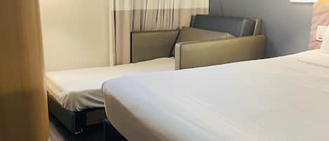 A hotel room with a large bed, a sofa, and a small table. There are curtains and a window with a view of the sky.