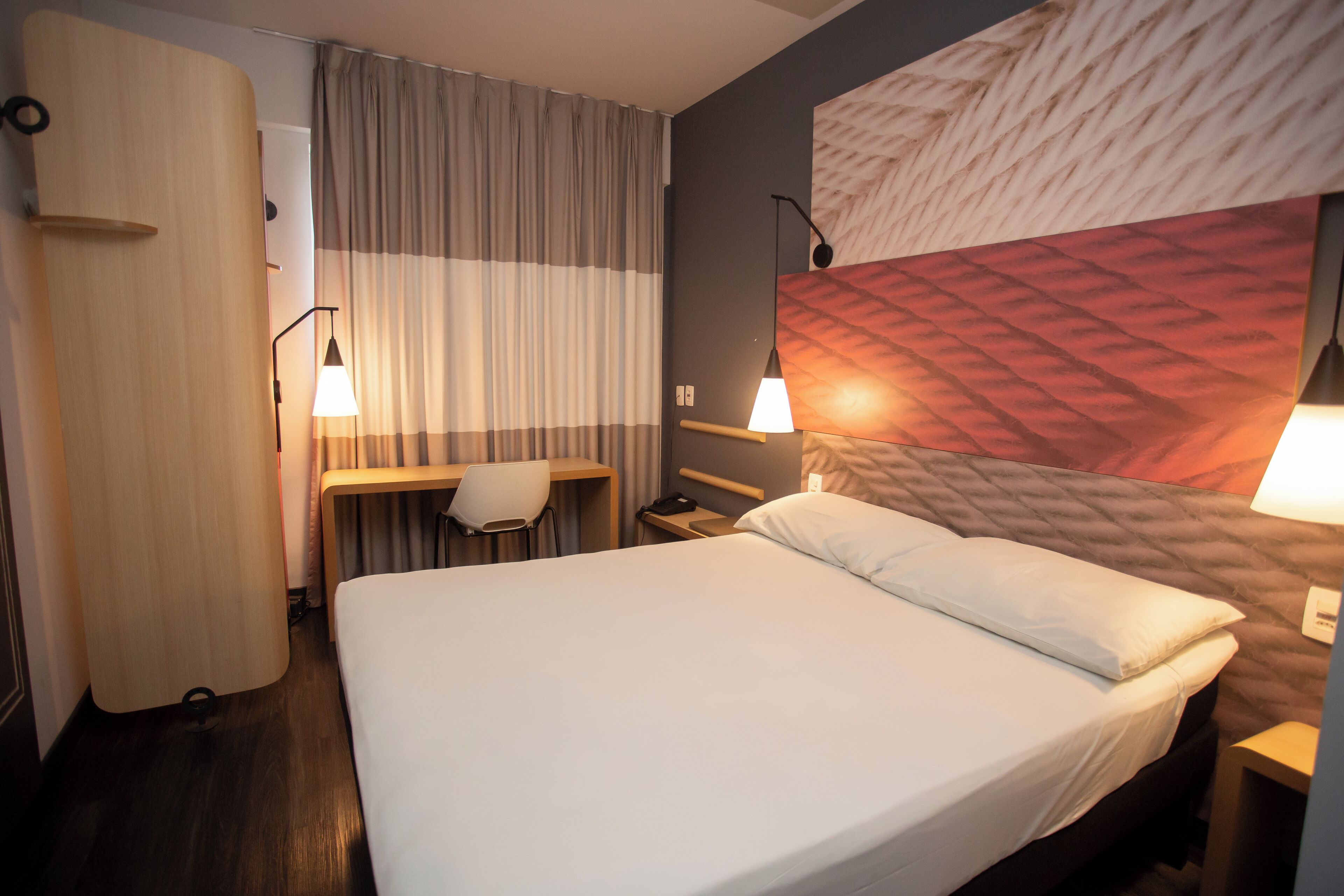 Een hotelkamer met een bed, een bureau, een stoel, een lamp en een gordijn.