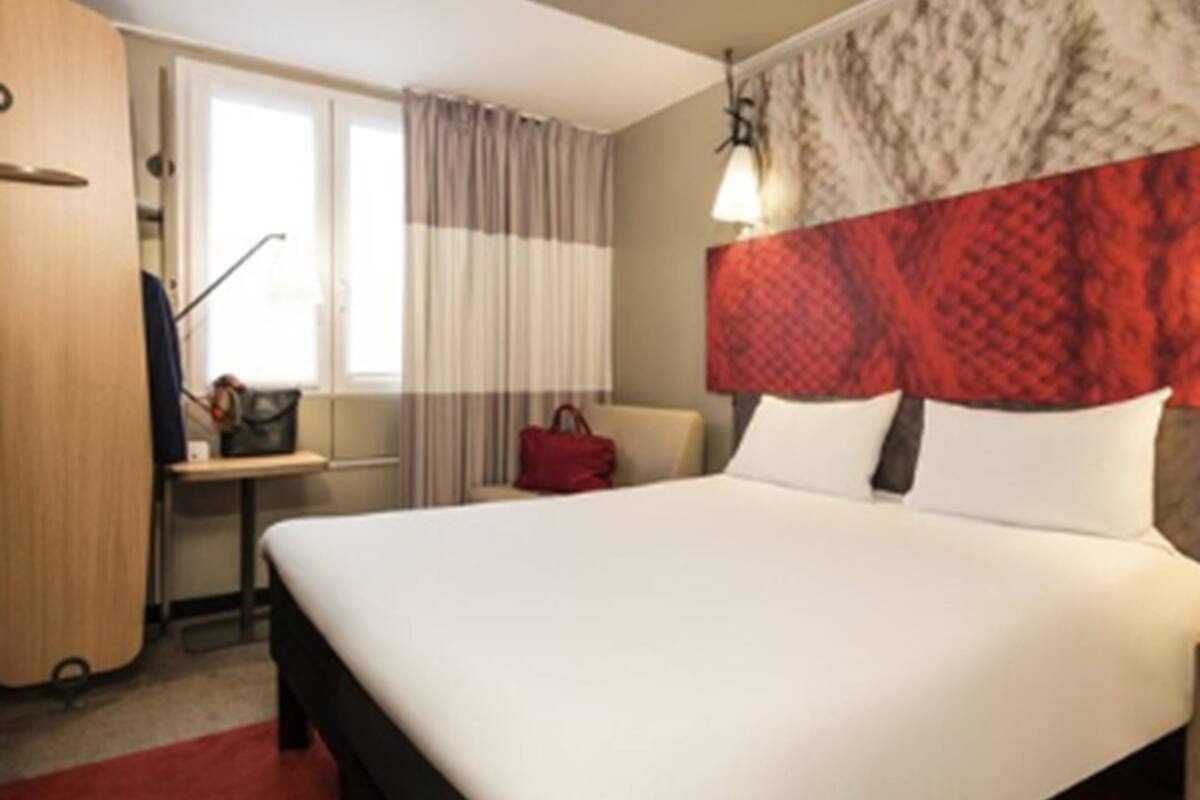 Una habitación de hotel con cama, escritorio, silla, armario y ventana con cortinas.