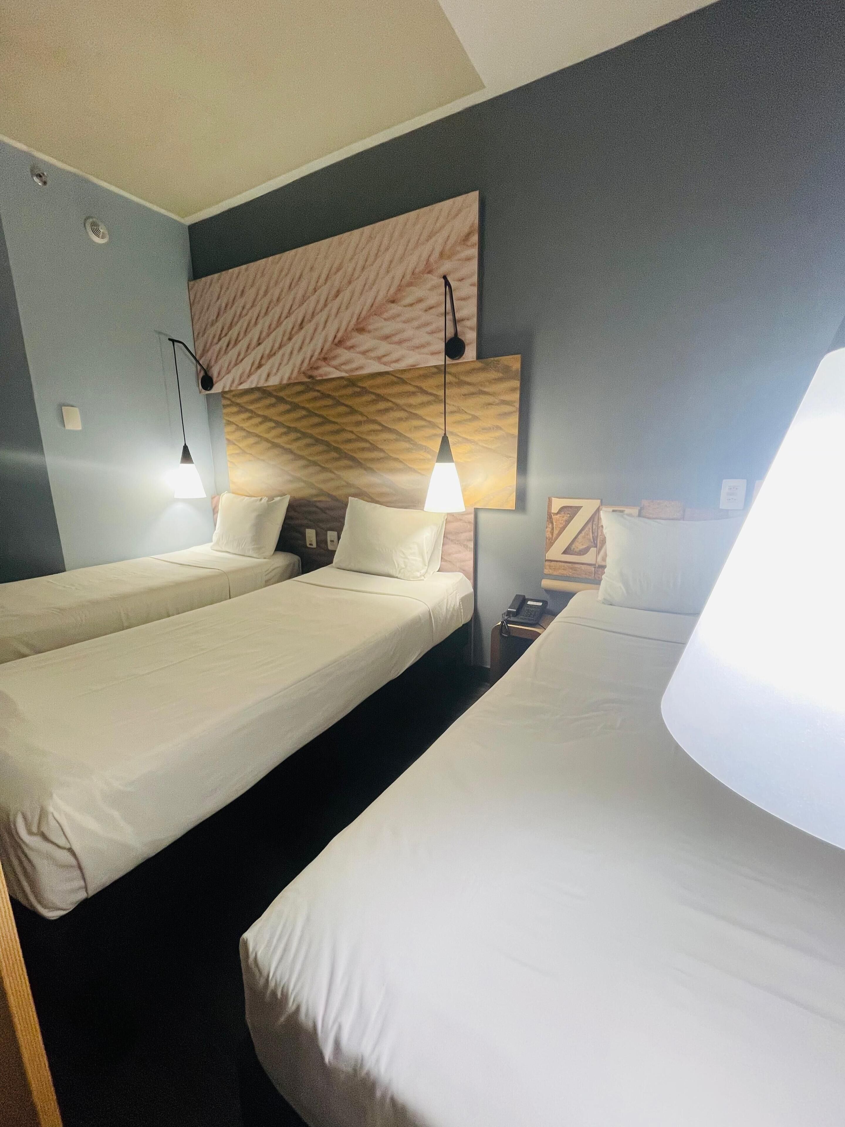 A hotel room with three single beds, each with a white mattress and pillow. There are two bedside lamps, one on each bed. The room has a modern design with a patterned headboard above the beds and a dark-colored wall.