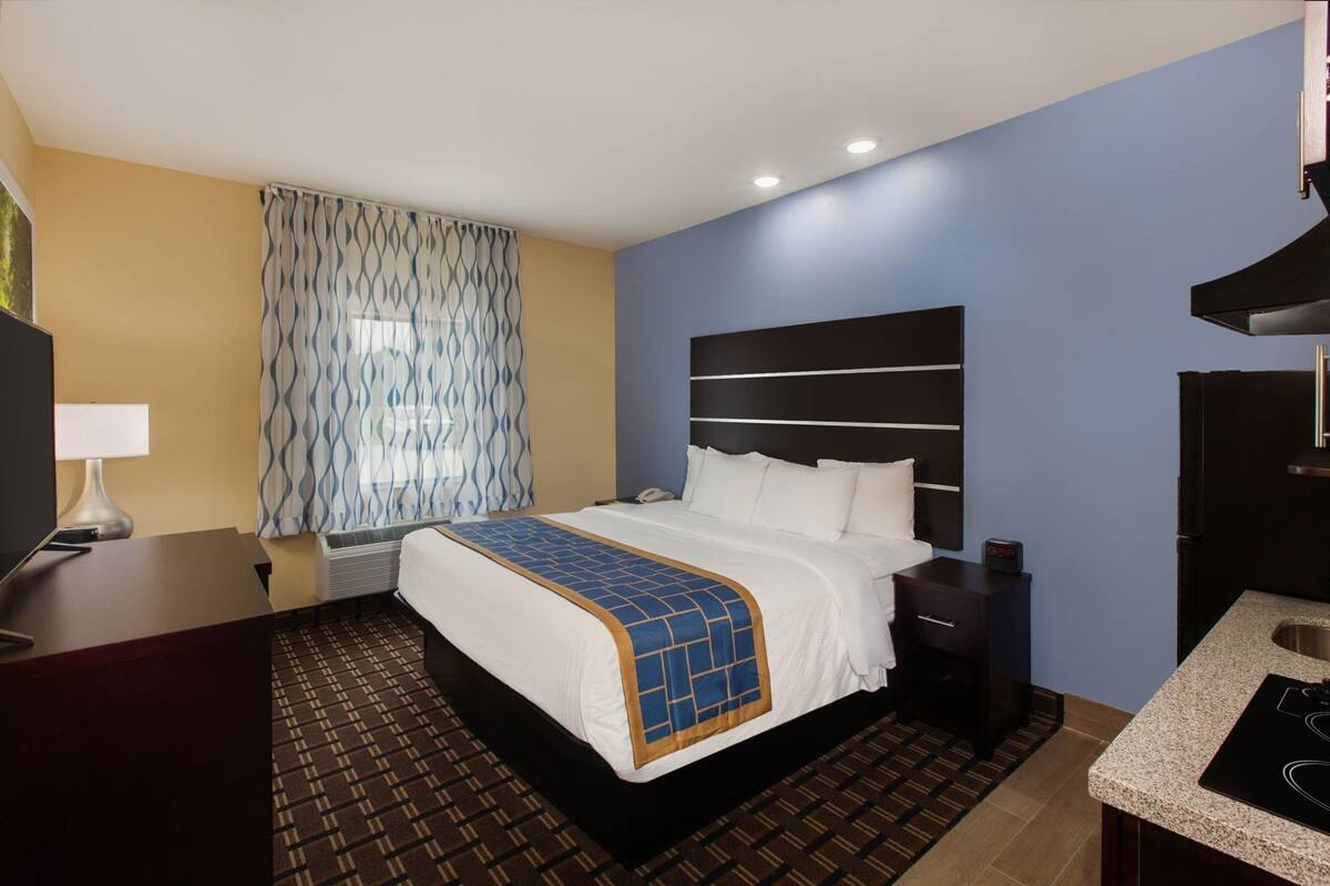 A hotel room with a bed, bedside table, desk, and a kitchenette.
