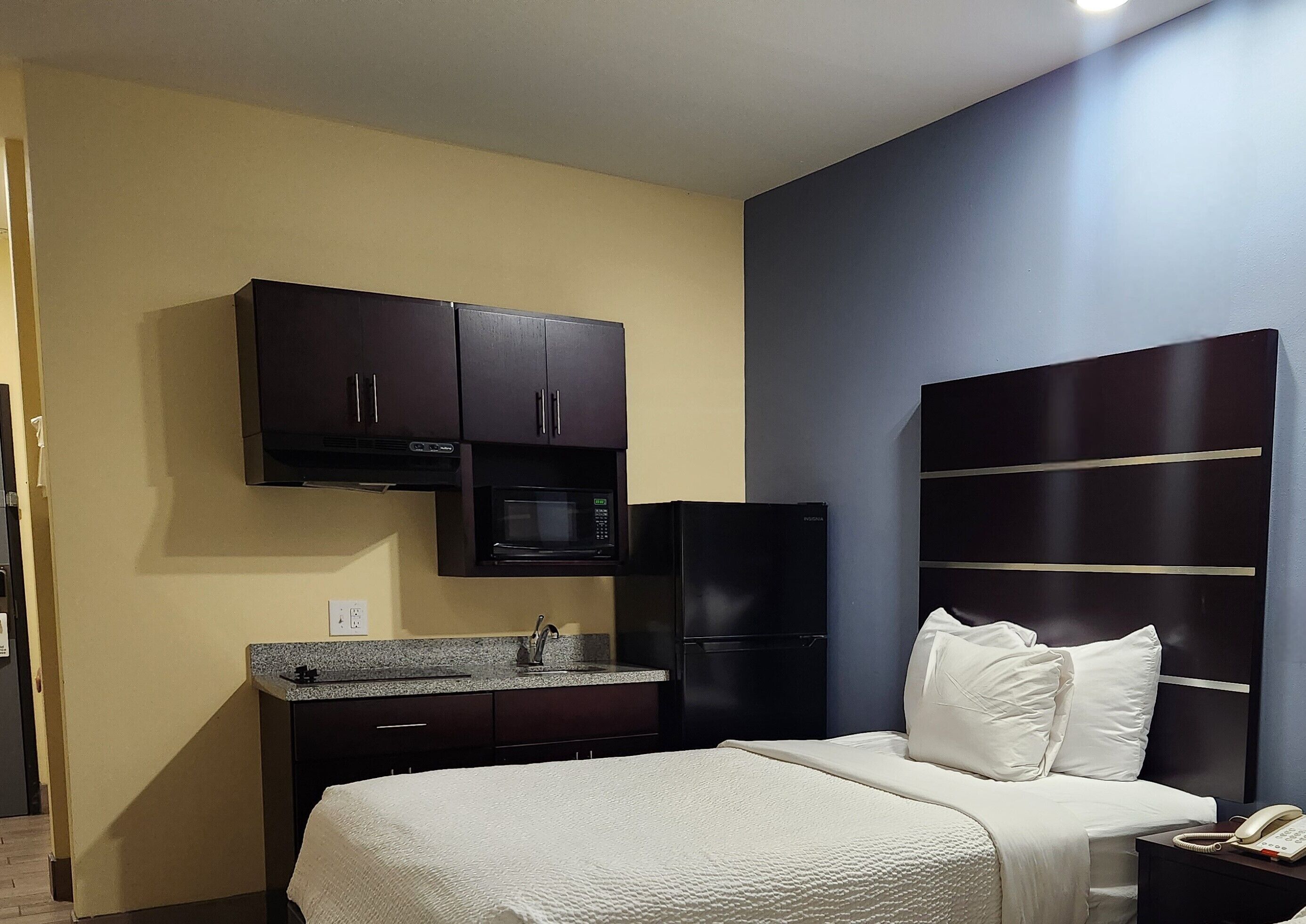 A compact hotel room with a bed, a small kitchenette, and a dark-colored headboard.