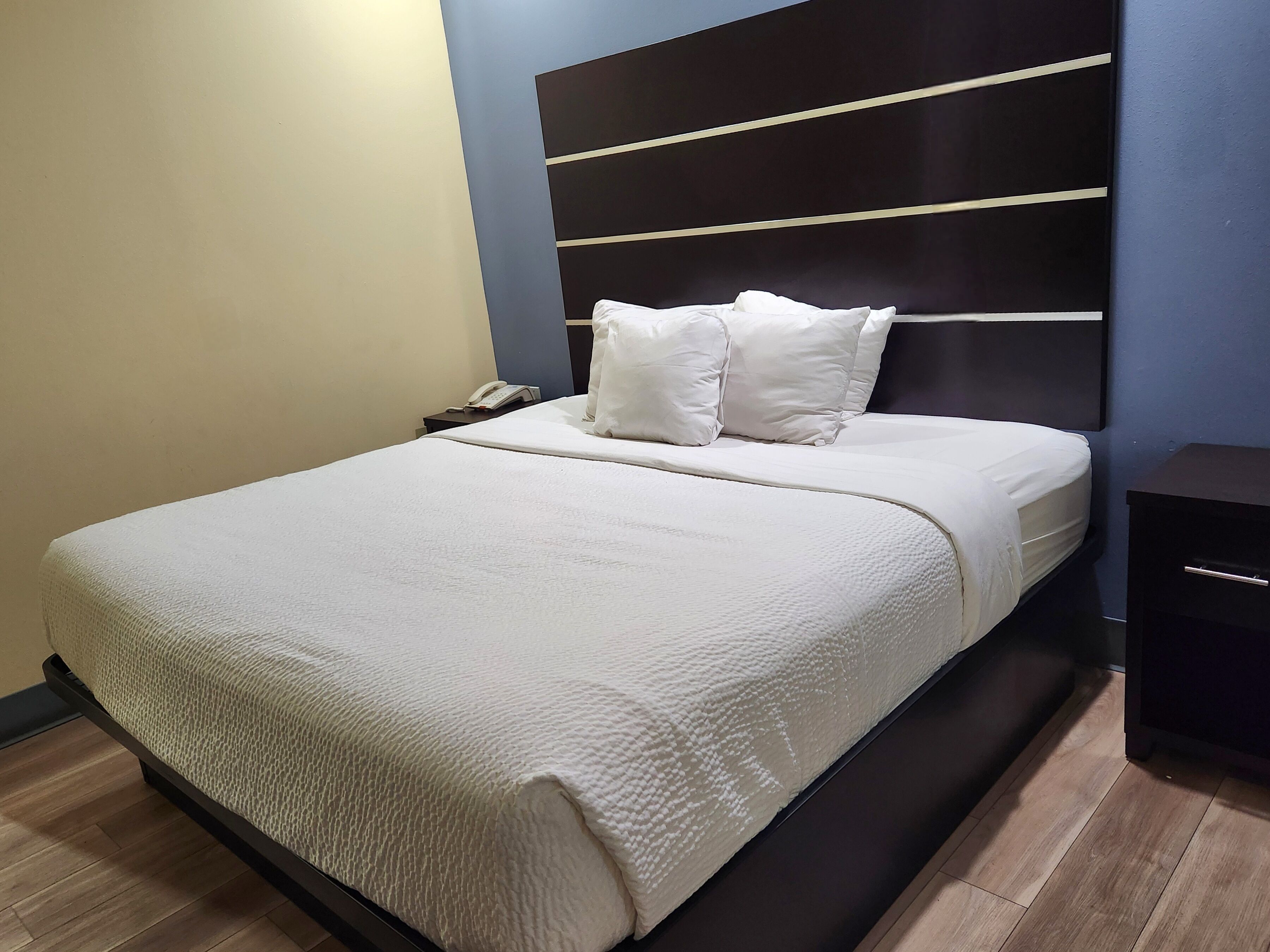A neatly made bed with a white comforter and pillows, a bedside phone, and a dark nightstand with a drawer.