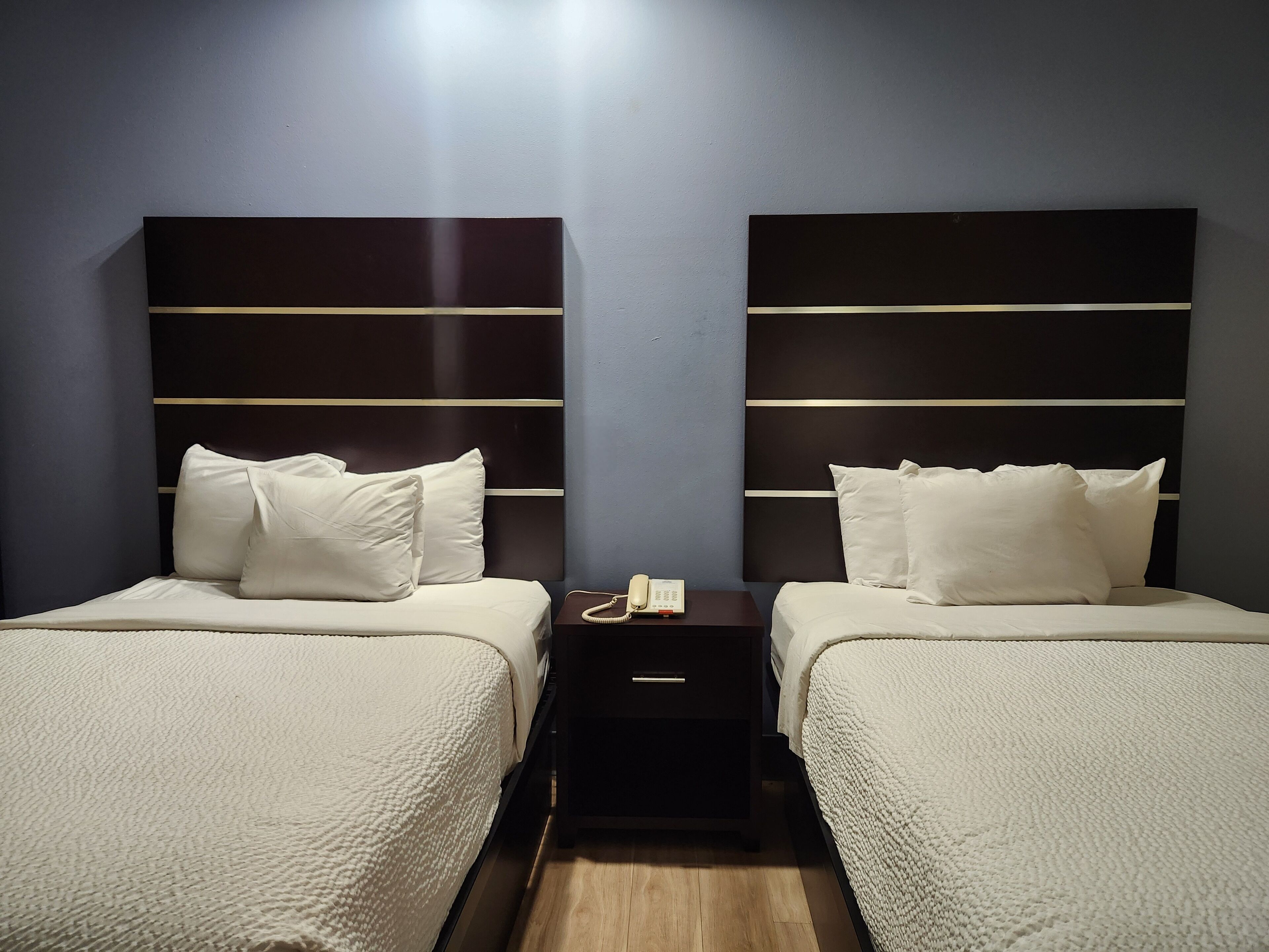 A hotel room with two beds, each with a white bedspread and pillows, and a dark headboard with horizontal lines. There is a small bedside table with a phone and a lamp.