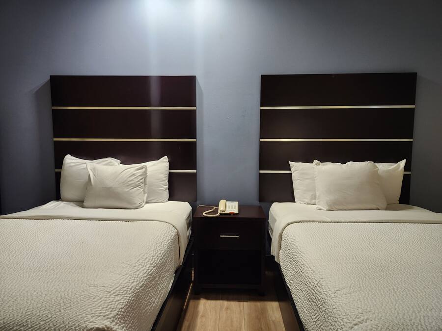 A hotel room with two beds, each with a white bedspread and pillows, and a dark headboard with horizontal lines. There is a small bedside table with a phone and a lamp.