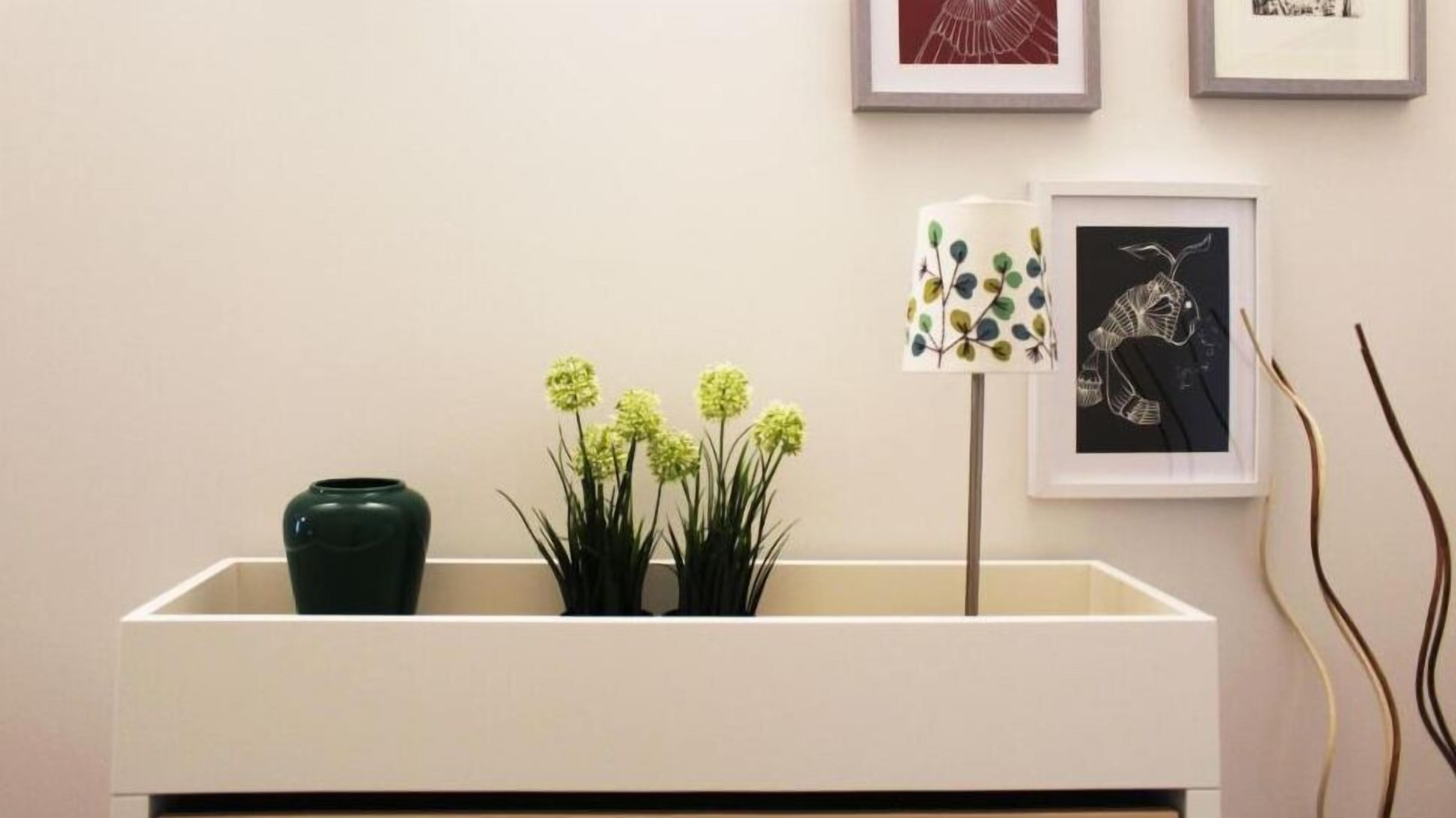 Una habitación minimalista con una consola blanca, un jarrón verde y una planta en maceta con flores amarillas. Hay dos obras de arte enmarcadas en la pared.
