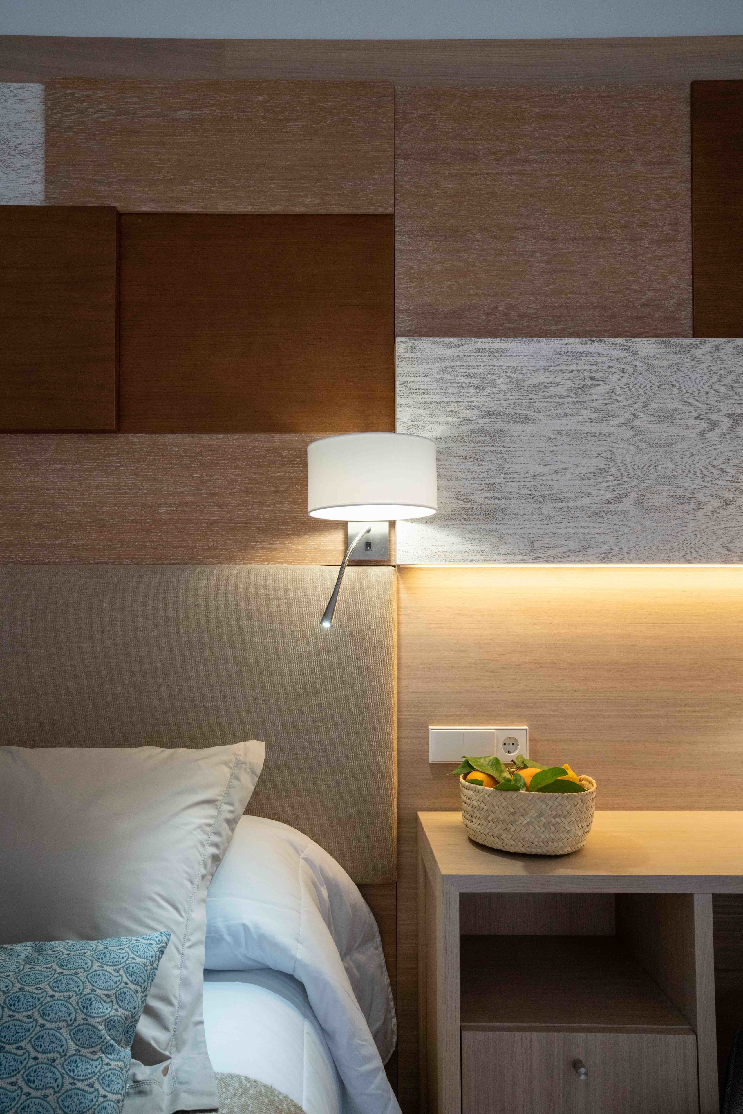A neatly made bed with a blue patterned pillow, a bedside table with a lamp, and a basket of fruit on the nightstand.