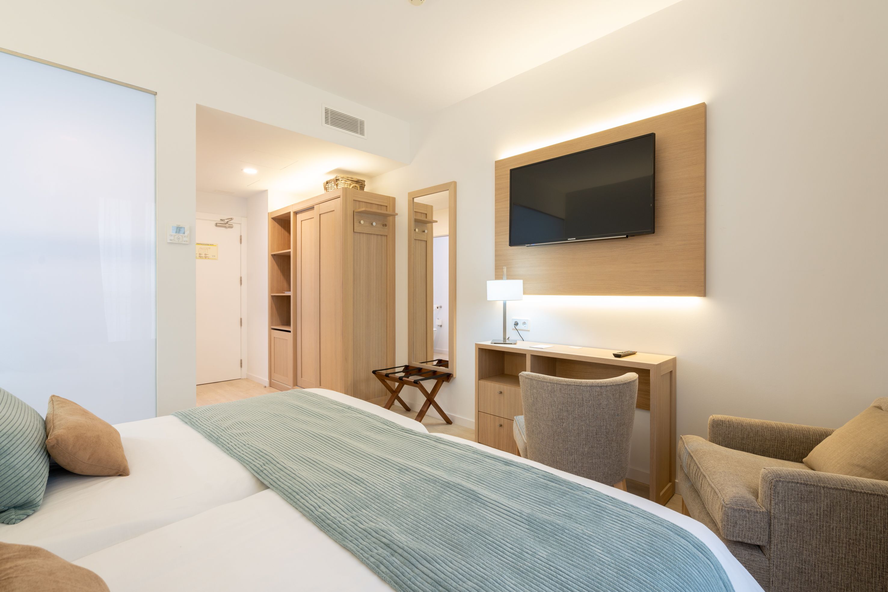A hotel room with a bed, a desk, a chair, a TV mounted on the wall, and a wardrobe.