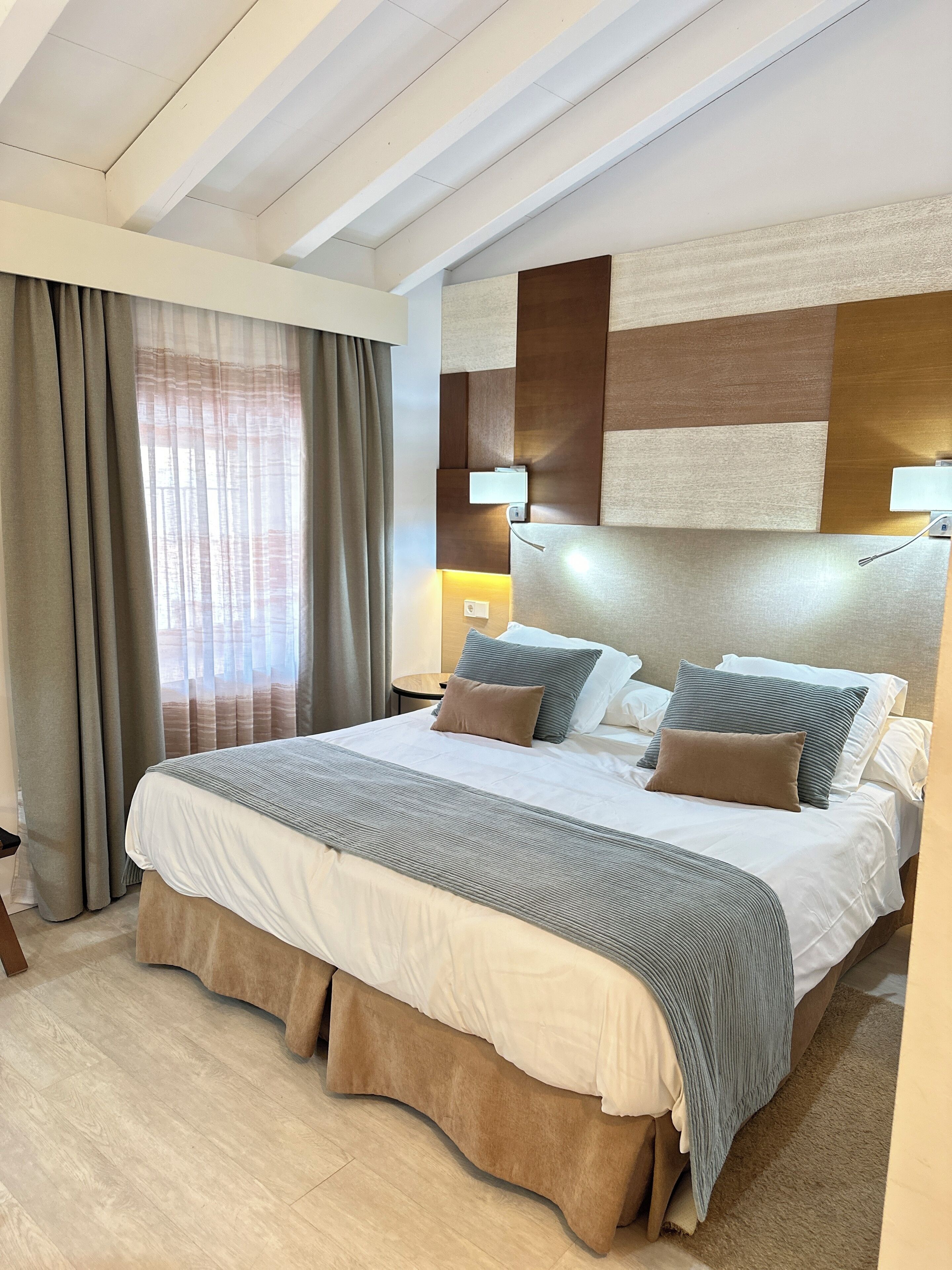 A hotel room with a large bed, a headboard with a geometric design, a bedside table with a lamp, and a view of the outside through sheer curtains.