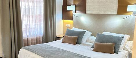 A hotel room with a large bed, a headboard with a geometric design, a bedside table with a lamp, and a view of the outside through sheer curtains.
