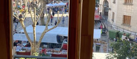 A view from a window overlooking a bustling square with market stalls, historic buildings, and mountains in the background.
