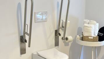 Een moderne badkamer met een wit toilet, twee wandbeugels, een witte prullenbak en een klein tafeltje met handdoeken en een haardroger.