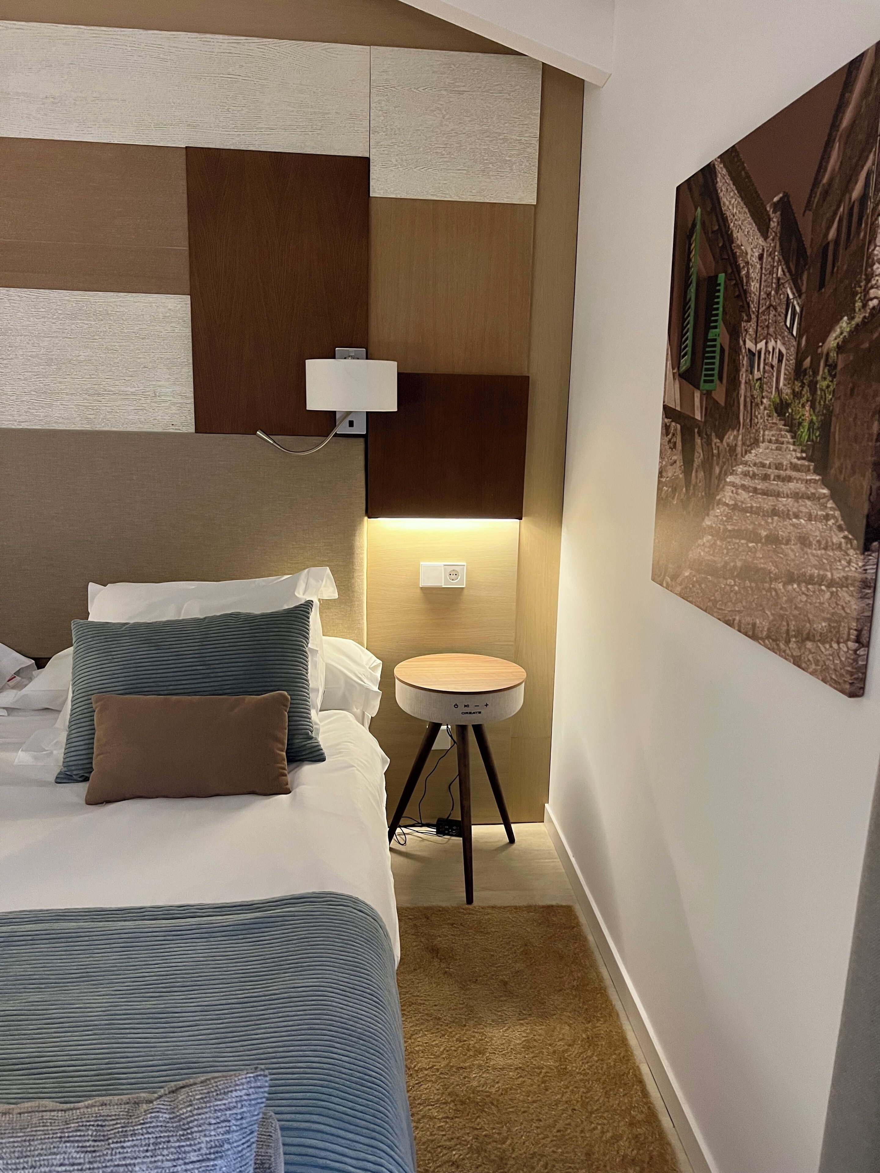 Une chambre d’hôtel avec un lit, une table de chevet, une lampe fixée au mur et un tableau représentant une scène de rue.