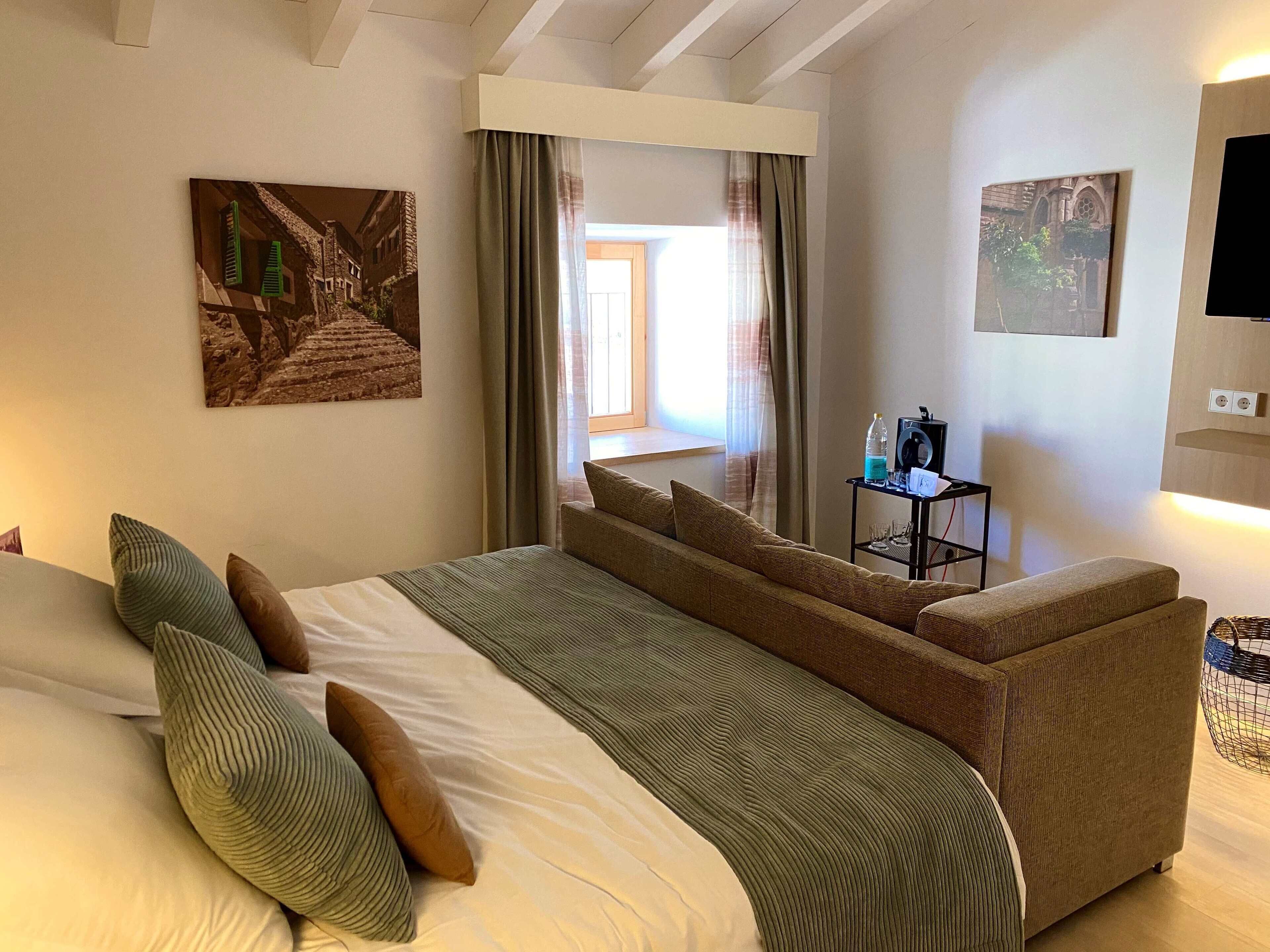 A hotel room with a bed, a sofa, a small table with a bottle and glasses, and two paintings on the walls.