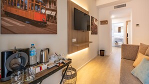 Una sala de estar moderna con un sofá, una mesa de centro con una tetera, una botella de agua y vasos. Un televisor está montado en la pared, y se muestra un tranvía en un cuadro.