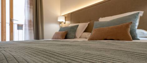 A hotel room with a large bed, a headboard with built-in lighting, and a view through a window with curtains.