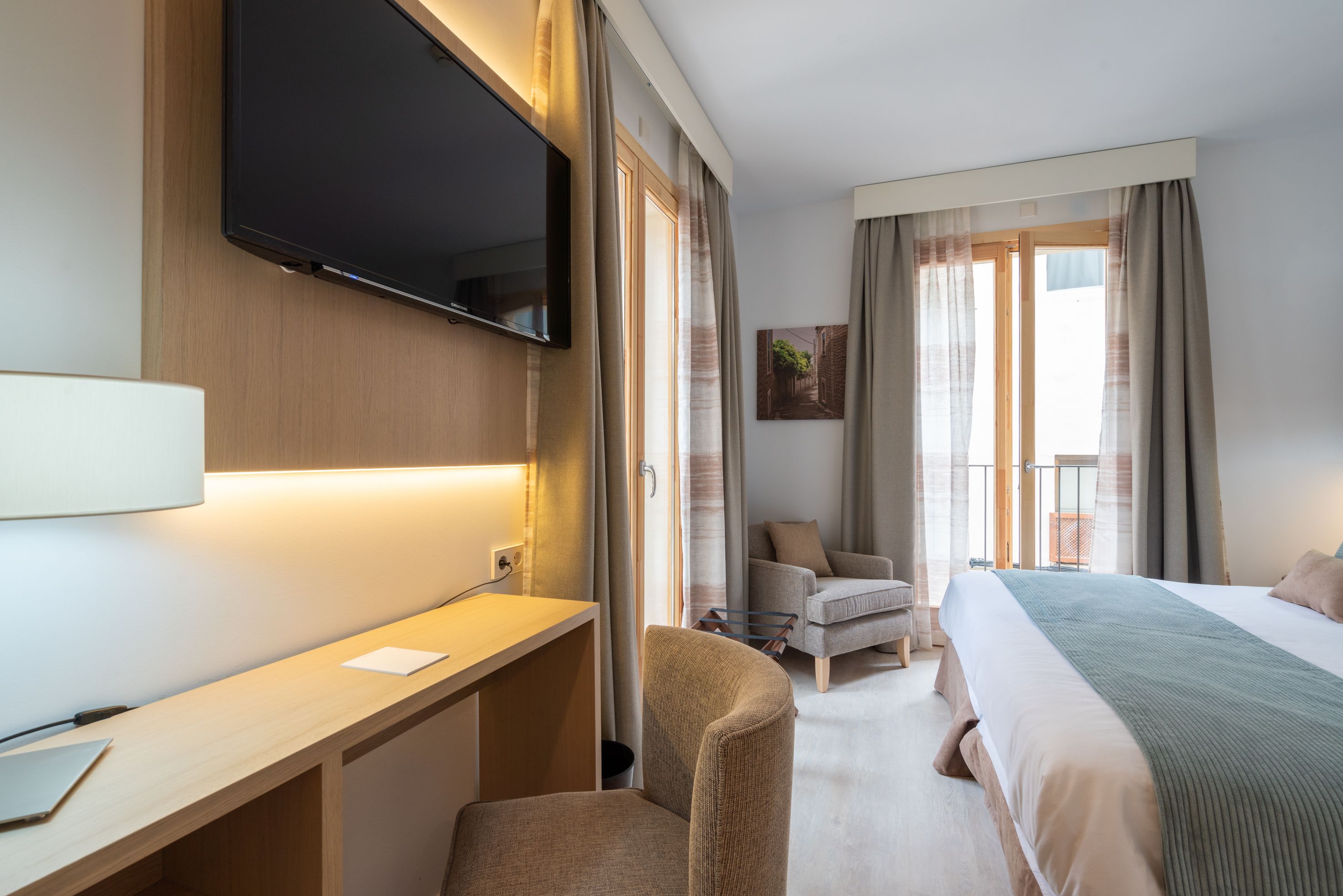 Ein Hotelzimmer mit Bett, Schreibtisch, Stuhl, Fernseher und einem Balkon mit Vorhängen.