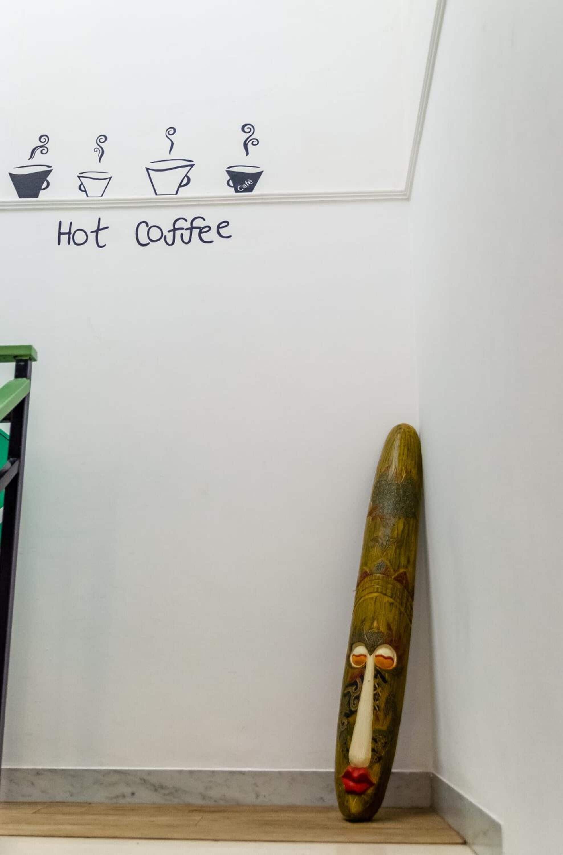 A wall with a drawing of four coffee cups and the text "Hot Coffee" below it. A wooden object with a face is leaning against the wall.