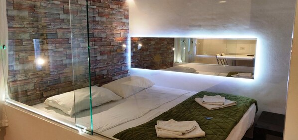 Une chambre d’hôtel moderne avec une cloison en verre, un mur au motif de briques, un lit avec du linge de maison blanc et une couverture matelassée verte.