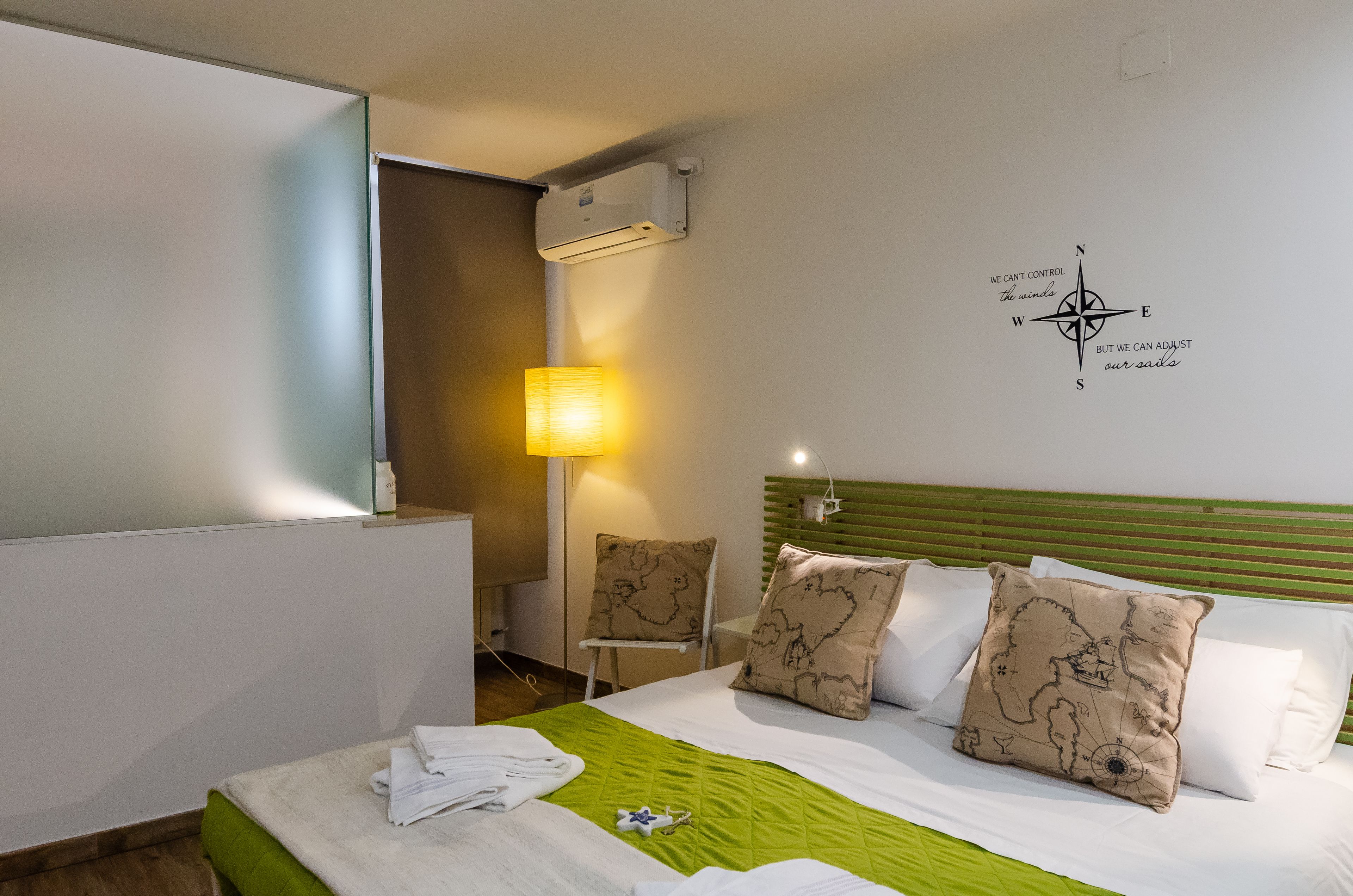 A hotel room with a bed, pillows, a green blanket, a wall-mounted air conditioner, a lamp, and a compass wall decoration.