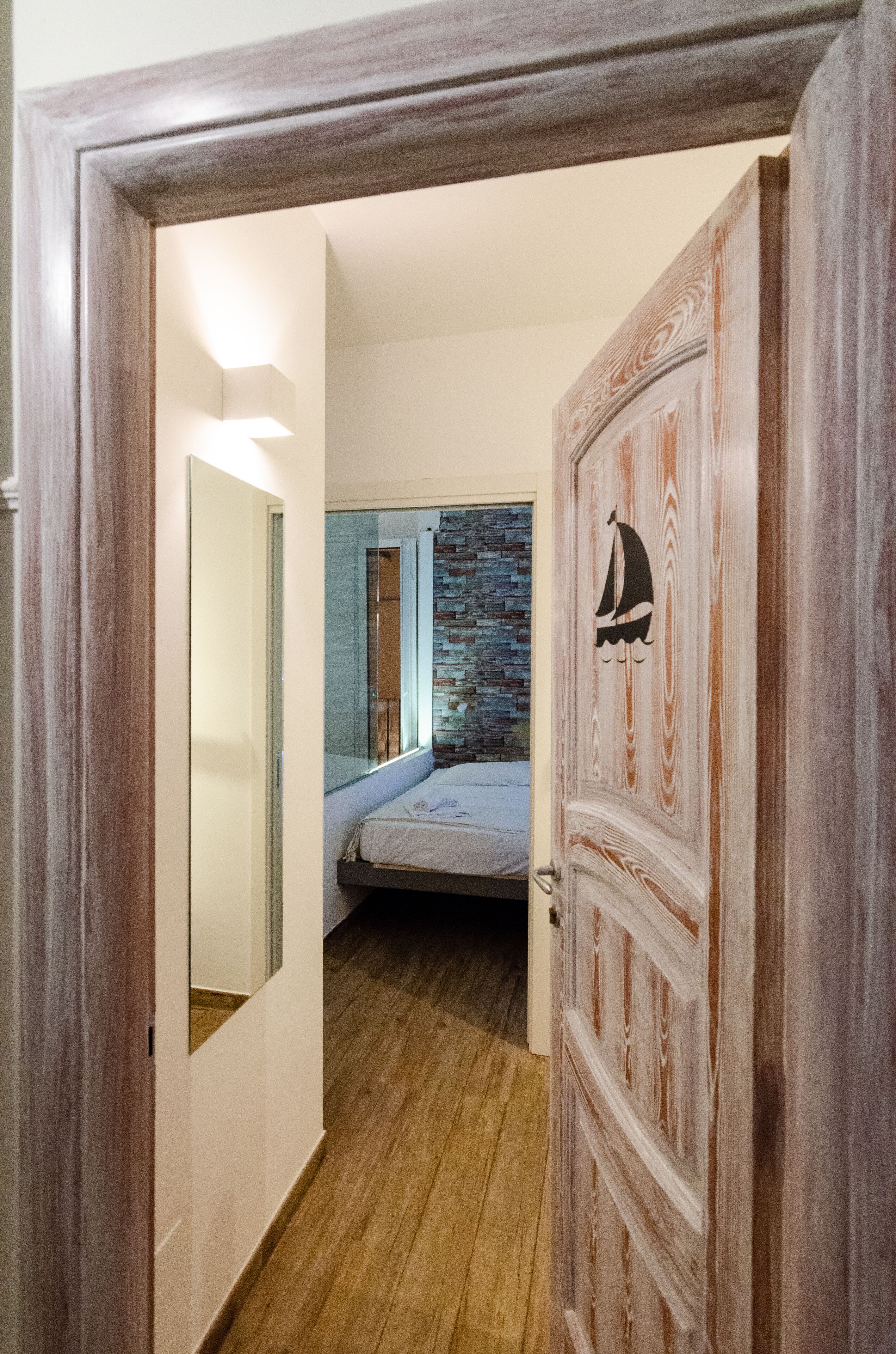 A hallway leading to a bedroom with a bed, a mirror, and a door with a sailboat design.
