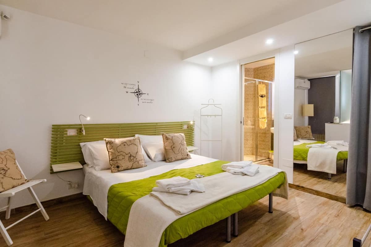 Habitación de hotel con cama, banca, silla, espejo y baño visible a través de una puerta abierta.