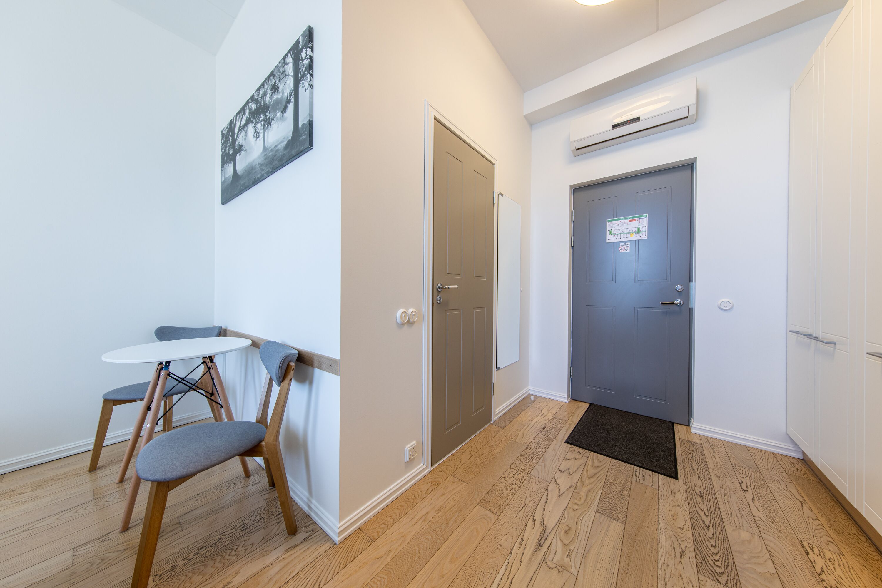 A room with a wooden floor, a white table with two chairs, a grey door, and a wall-mounted air conditioner.