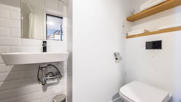A modern bathroom with a white sink, mirror, and toilet. There are wooden shelves above the toilet holding towels.