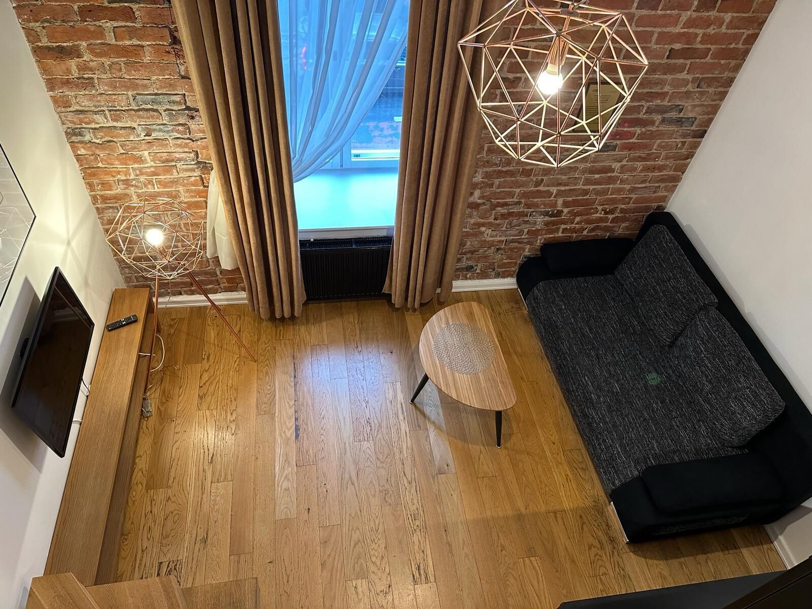 Un salon moderne avec un plancher en bois, un canapé noir, un luminaire suspendu aux motifs géométriques et une vue sur une piscine à travers de grandes fenêtres.