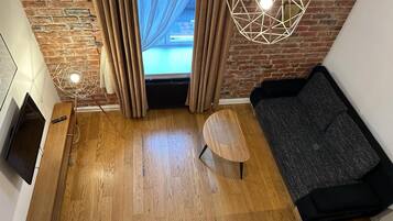 Un salon moderne avec un plancher en bois, un canapé noir, un luminaire suspendu aux motifs géométriques et une vue sur une piscine à travers de grandes fenêtres.