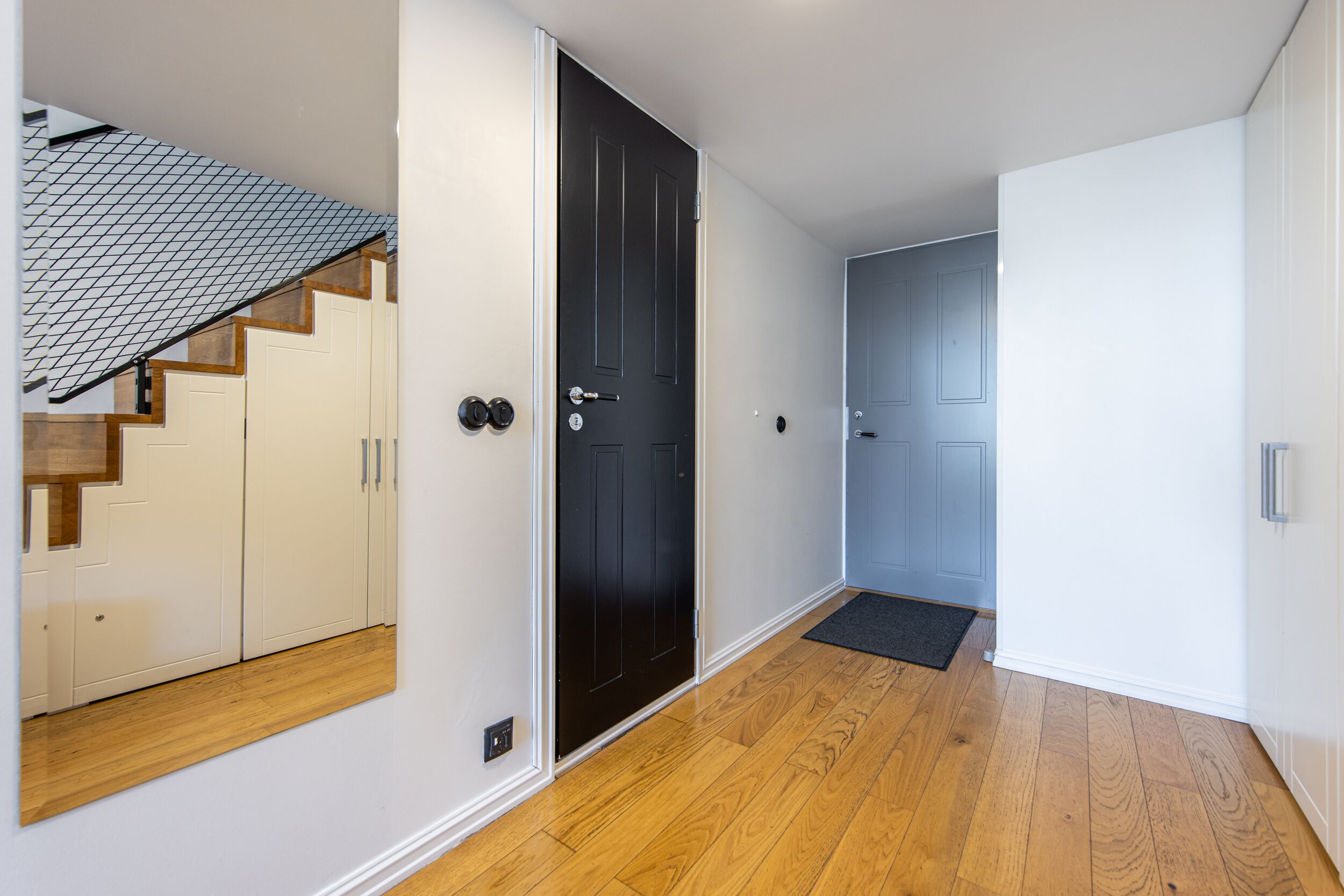 Un pasillo con suelo de madera, una puerta negra y una escalera con paredes color crema y escalones de madera.