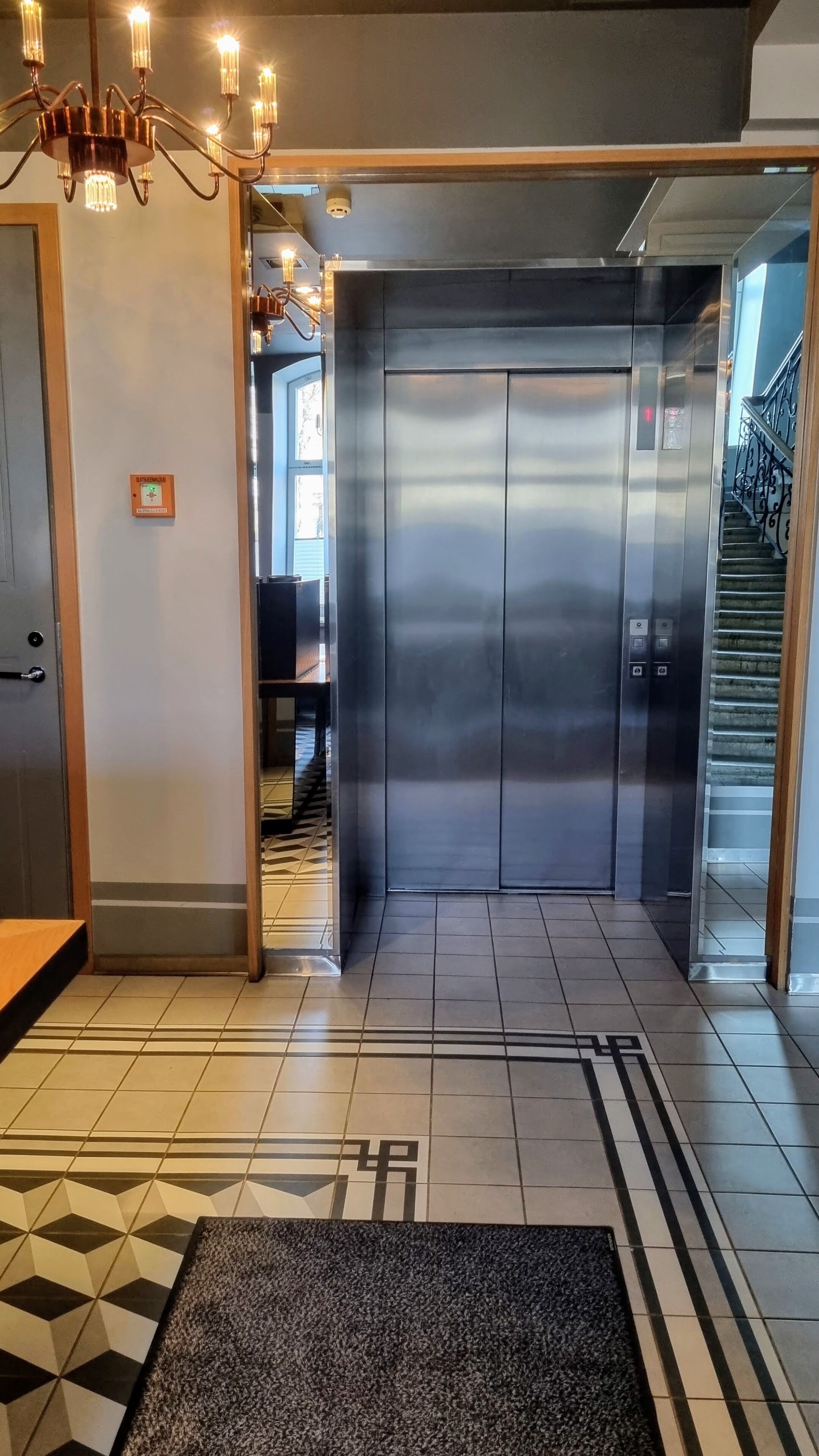 Un ascensor con detalles metálicos, un suelo a cuadros blanco y negro, y una araña colgante en la parte superior.
