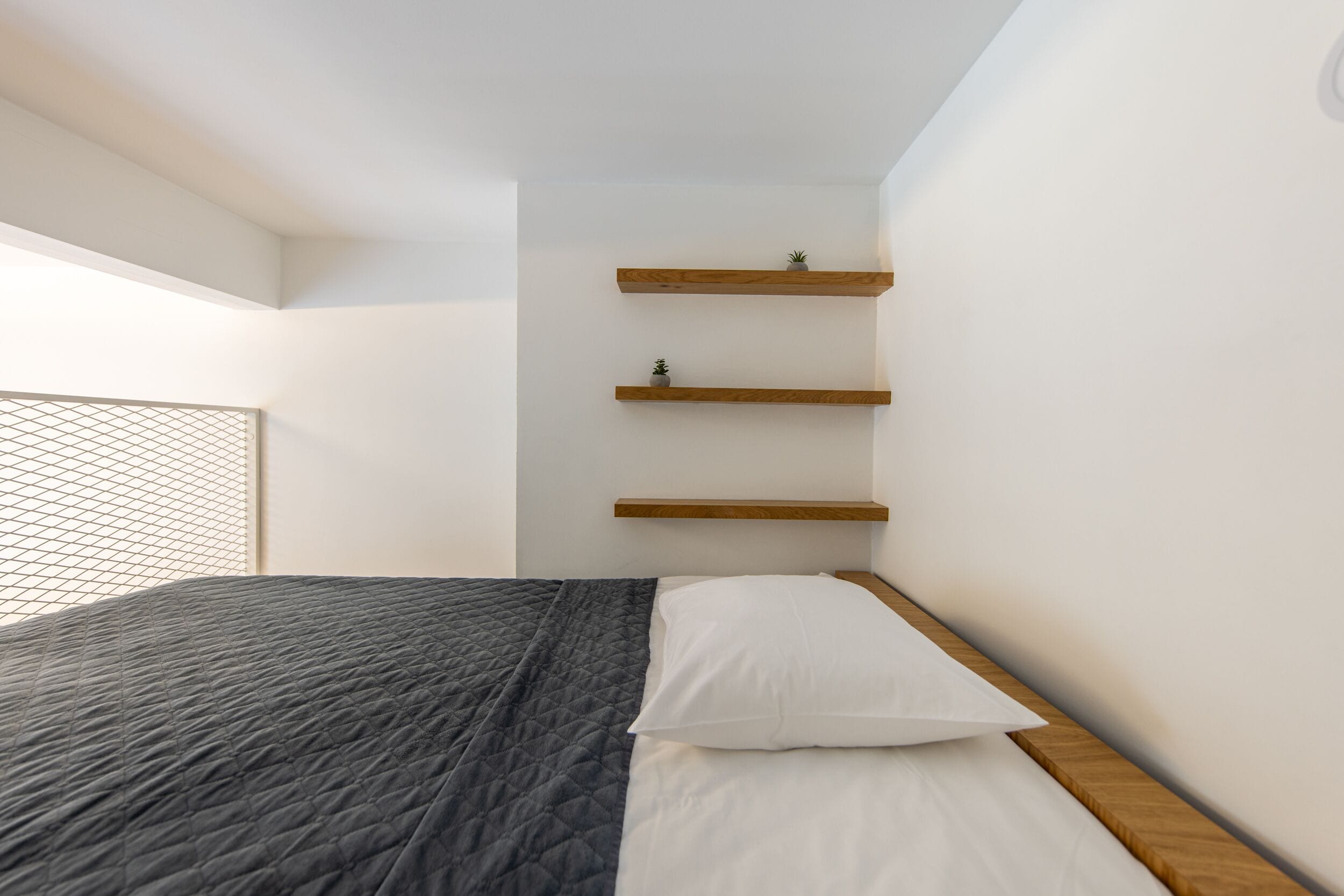 A bedroom with a bed, white bedding, a dark quilted blanket, and three wooden shelves against the wall.