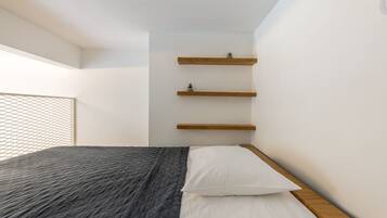 A bedroom with a bed, white bedding, a dark quilted blanket, and three wooden shelves against the wall.