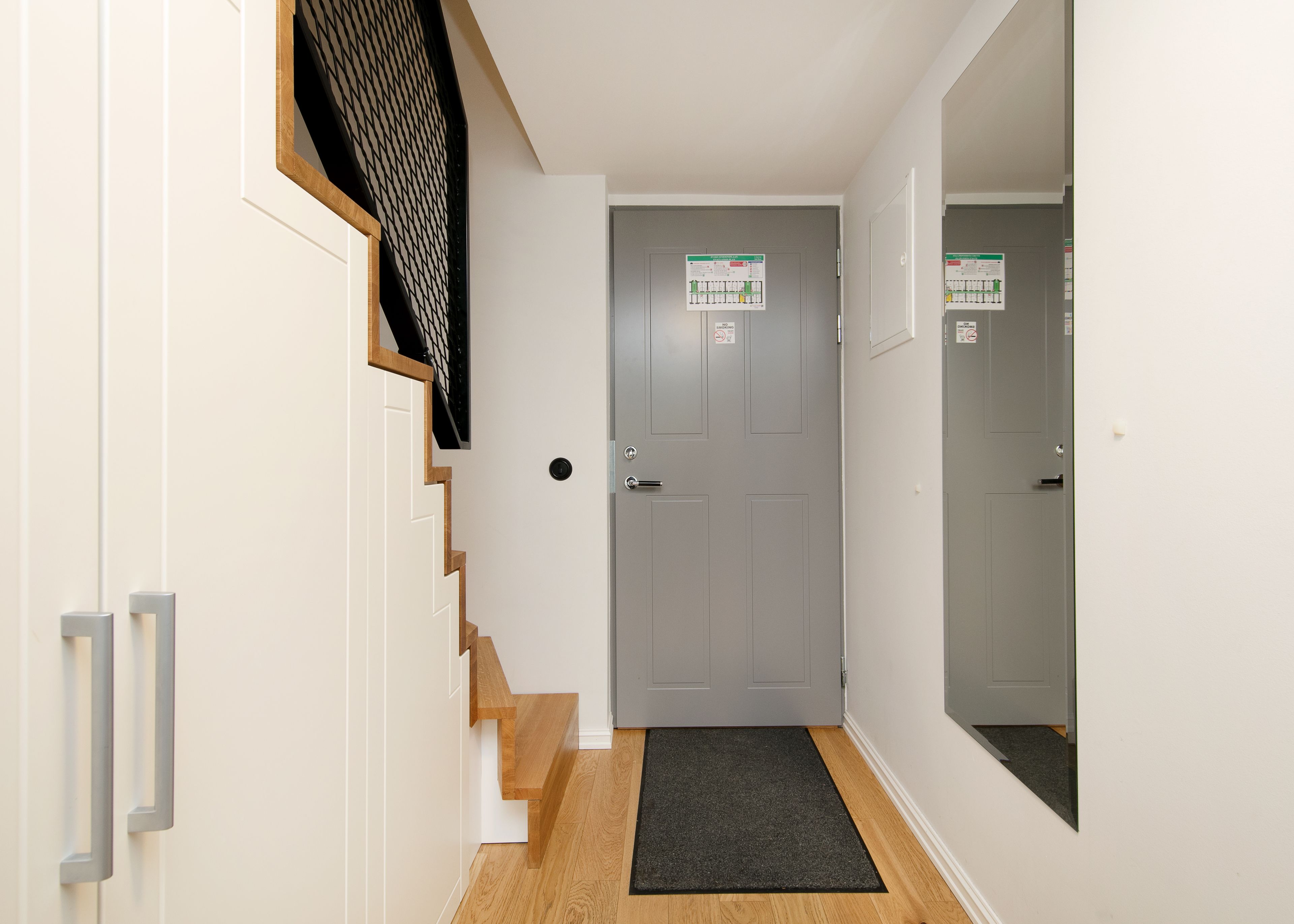 A hallway with a grey door, a black mat at the entrance, and a staircase with wooden steps.