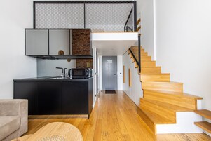 A modern kitchen with a black countertop, a microwave, and a brick wall. A wooden staircase leads to a door.
