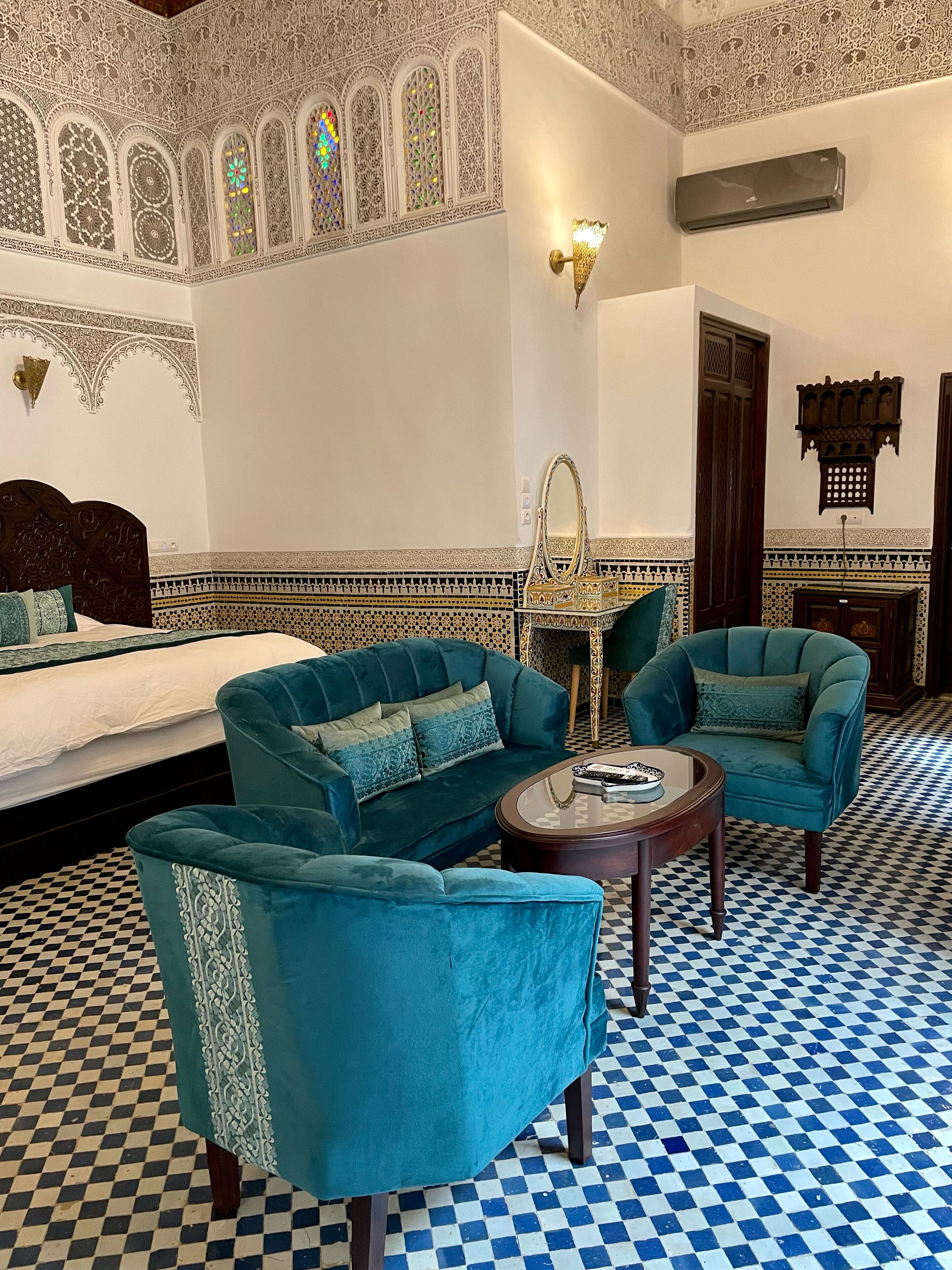 A room with a checkered floor, a bed, two teal armchairs, a small round table, and a wall with intricate patterns and a mirror.