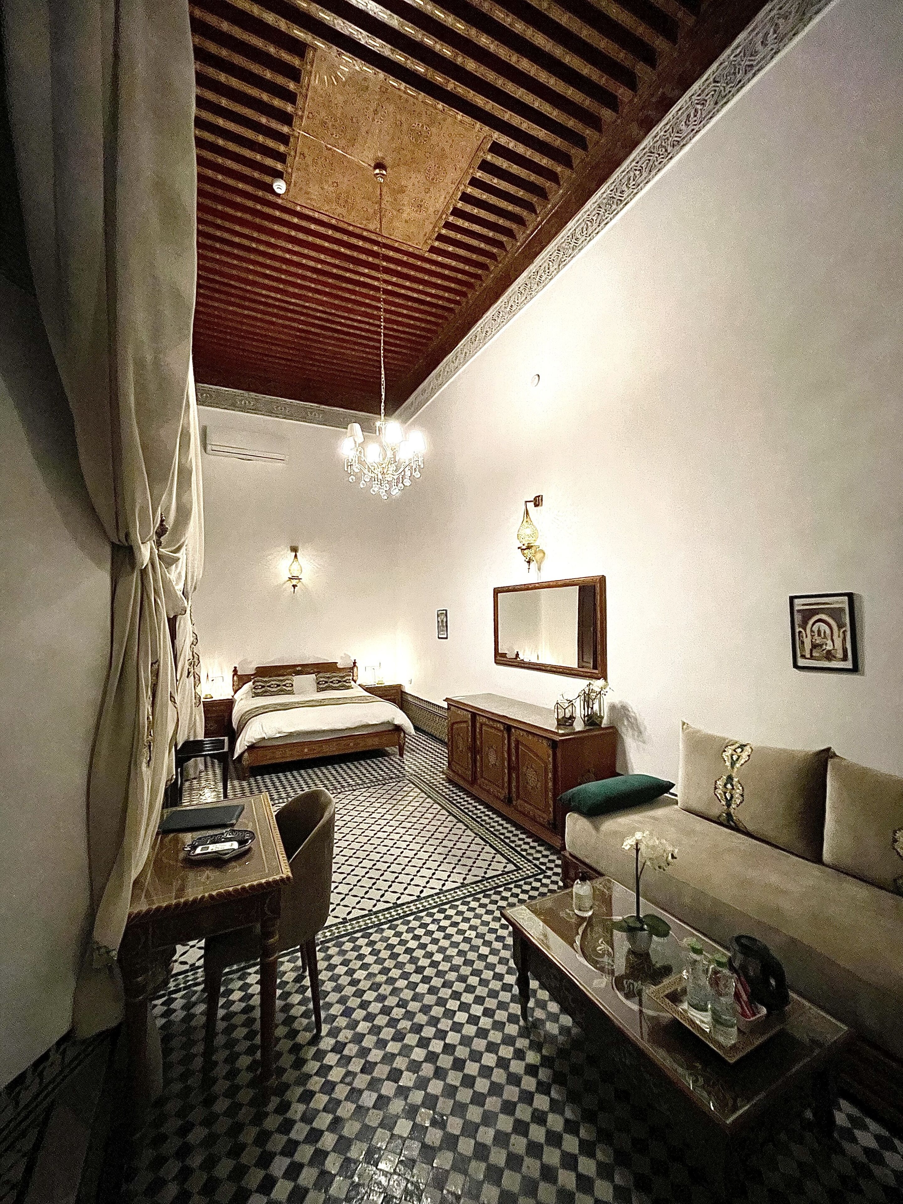 A hotel room with a checkered floor, a bed, a sofa, a dining table, and a chandelier.