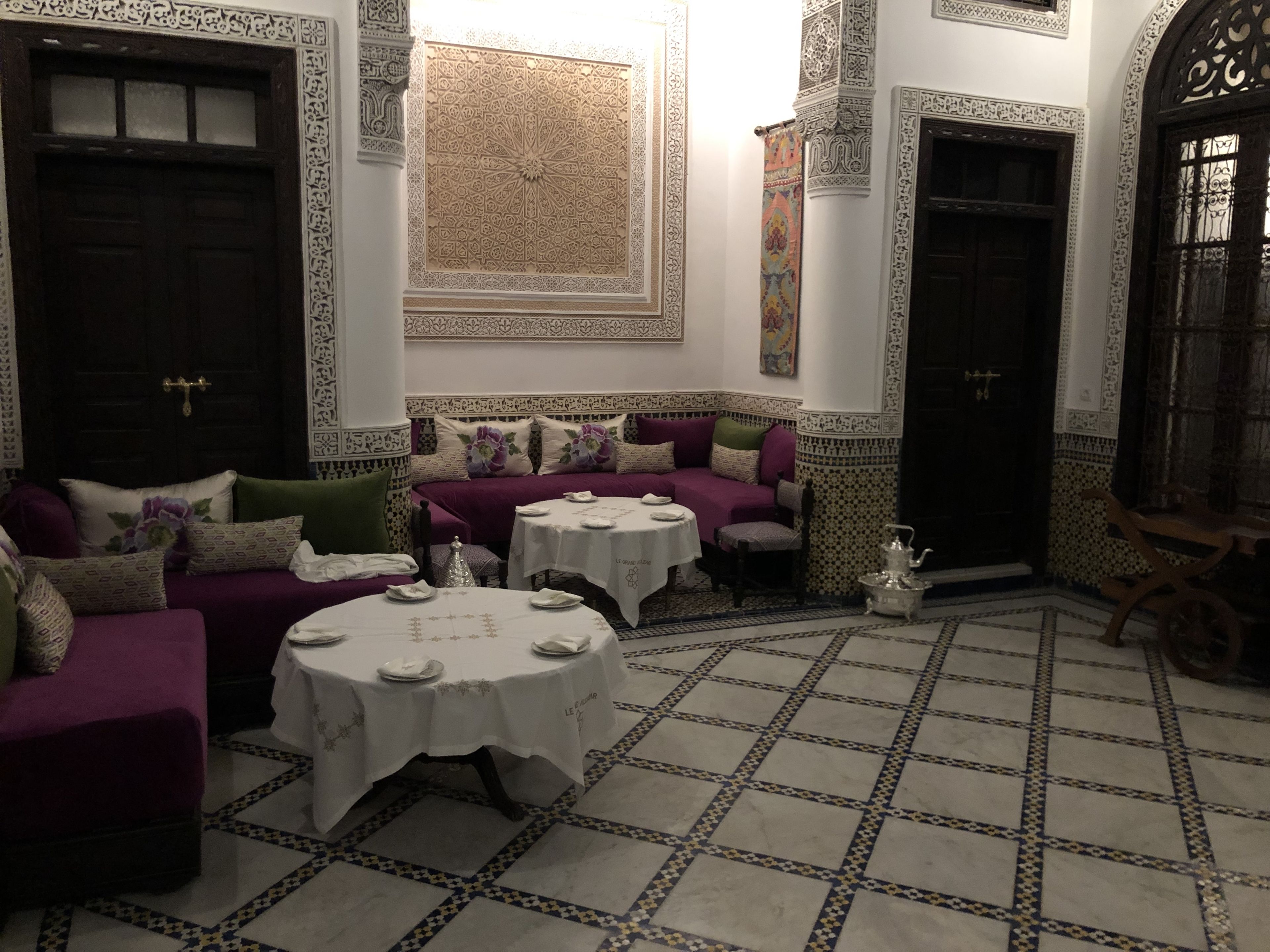Habitación en estilo marroquí con un sofá morado, dos mesas redondas con manteles blancos, suelo de baldosas estampadas y decoraciones ornamentales en las paredes.
