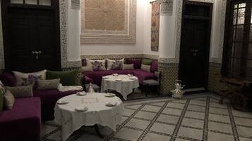 Habitación en estilo marroquí con un sofá morado, dos mesas redondas con manteles blancos, suelo de baldosas estampadas y decoraciones ornamentales en las paredes.
