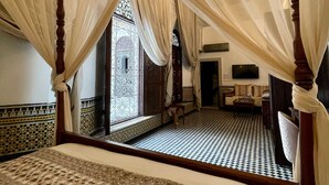 Un dormitorio con cama con dosel, alfombra estampada y vista a un patio a través de una ventana con celosía detallada.