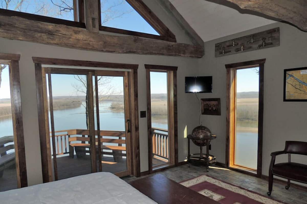 A bedroom with a large bed, wooden floor, a desk, a chair, a TV, and a view of a lake through the door.