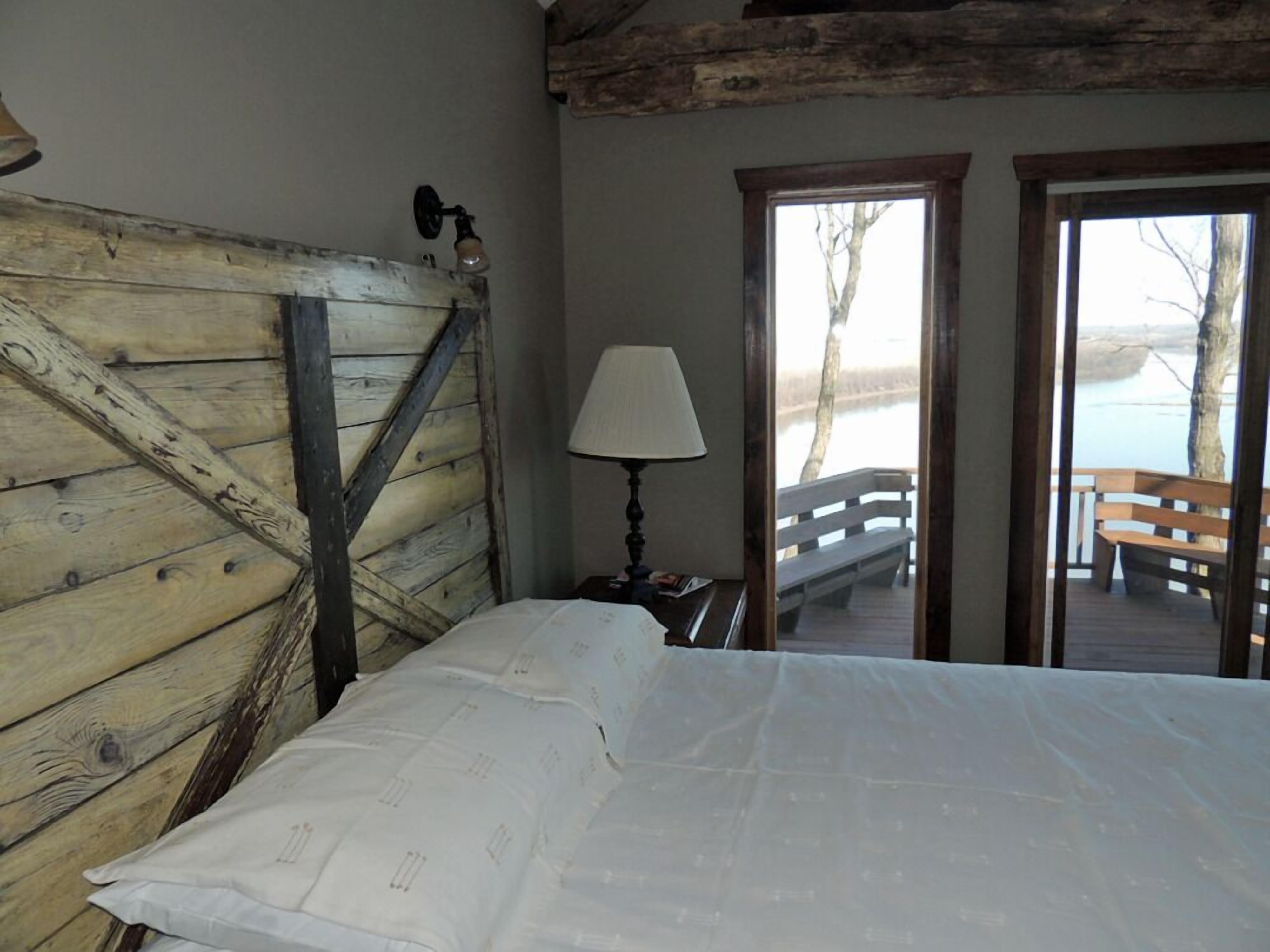 A rustic wooden room with a bed, a lamp, and a view of a wooden deck and lake.