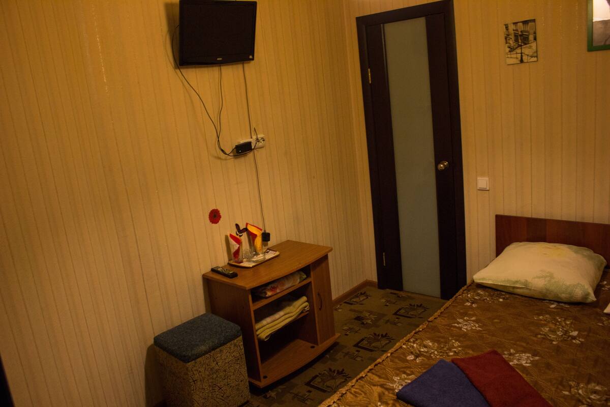 A room with a bed, a wooden nightstand, a television mounted on the wall, and a door.