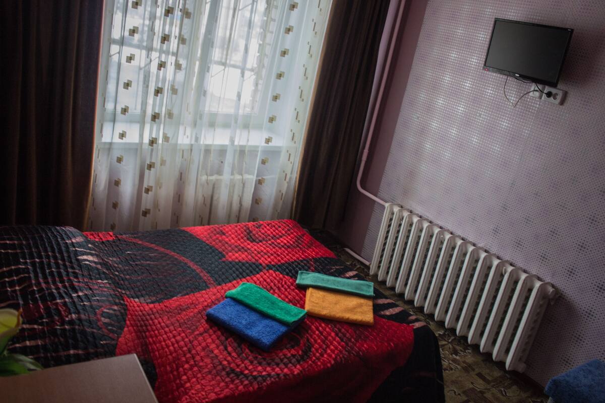 A hotel room with a bed, a radiator, a television mounted on the wall, and a window with curtains.
