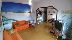 Una sala de estar con un cuadro grande de una playa, un sofá, una silla, un televisor y una zona de cocina con estanterías.