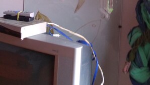Una habitación con una pizarra que muestra la palabra 'GRATIDAD' y dibujos de pájaros. Hay un televisor sobre un pedestal con un control remoto y un decodificador.