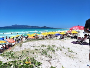 A beach with people sunbathing and swimming, colorful umbrellas, and a clear blue sky. - Ferias do Massari (Cabo Frio)
